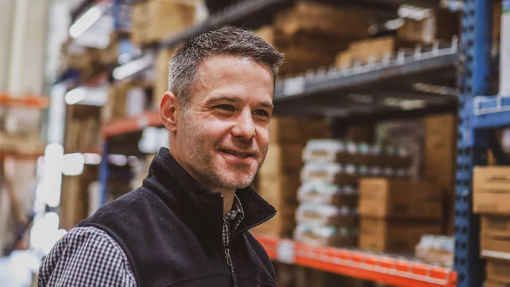 Warehouse worker wearing a black vest standing in a well-organized storage area with shelves stocked with boxes and medical supplies